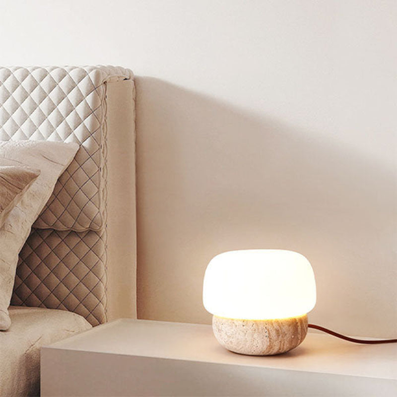 A small, glowing table lamp with a rounded white shade and a textured, light-colored base sits on a light-colored nightstand. The lamp's brown cord extends to the right. The nightstand is next to a bed with a quilted headboard and soft pillows against a light beige wall. The lamp emits a warm light, illuminating the bedside area.