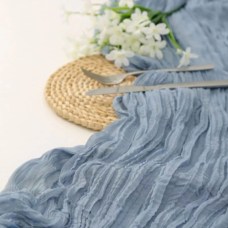 Soft, crinkled blue table runner with silver cutlery on a woven placemat, accented by white flowers.