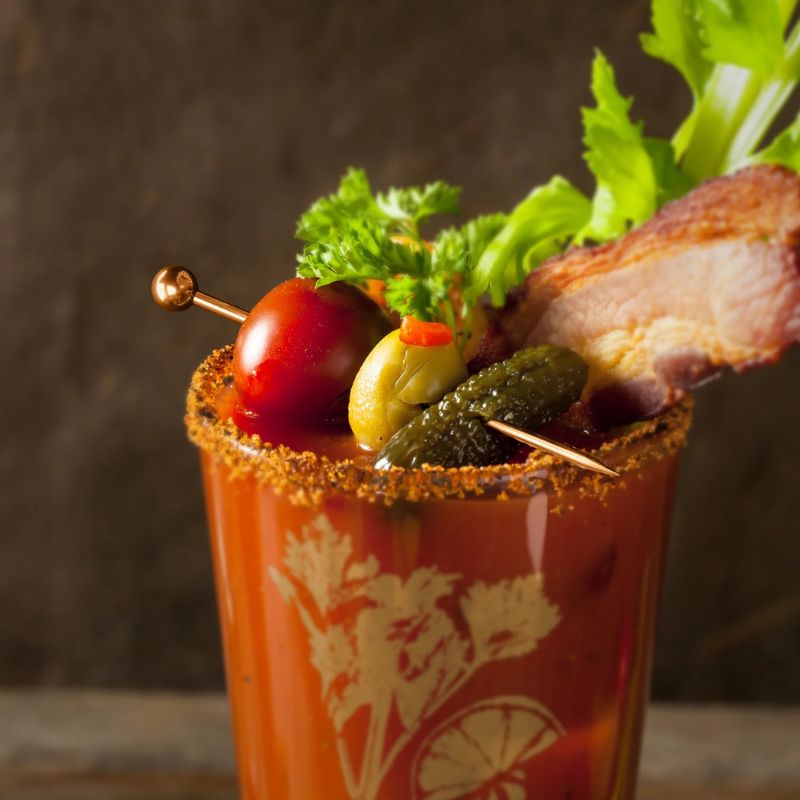 A Bloody Mary cocktail with garnishes, including bacon, tomatoes, olives, pickles, and celery.