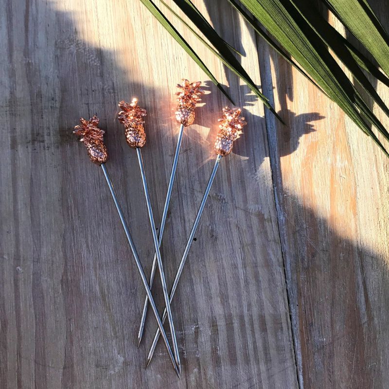 Four stainless steel cocktail picks with small, detailed gold pineapple-shaped toppers, arranged on a weathered wooden surface with the shadow of a palm leaf cast across them.