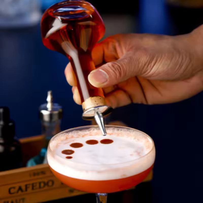 A hand uses a uniquely shaped glass bottle with a spout to add drops of liquid garnish to a frothy cocktail in a coupe glass. A wooden crate with bar tools is visible in the background.