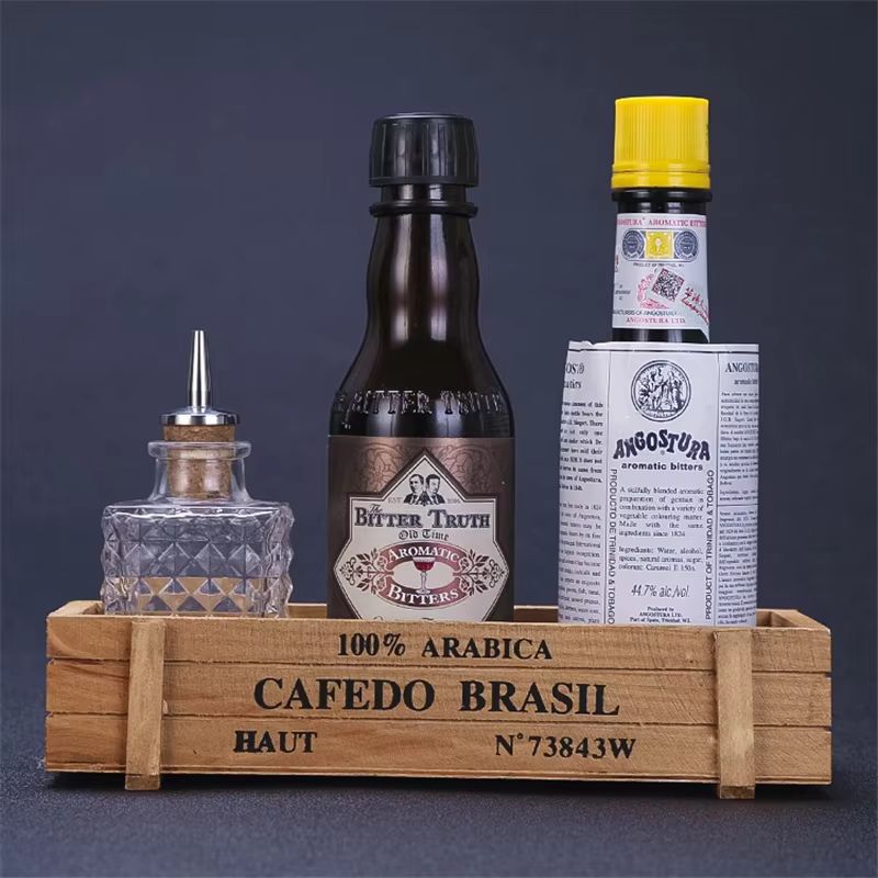 Wooden crate labeled "CAFEDO BRASIL" holding three bottles: a small glass bitters bottle with a pour spout, a dark brown bottle with vintage-style labeling, and a tall bottle wrapped in white paper with a yellow cap.