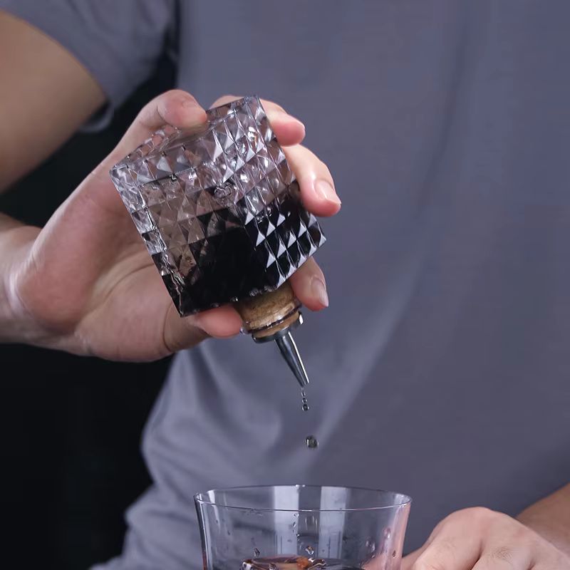 A person pours a dark liquid from an inverted, diamond-patterned glass bottle with a spout into a tumbler filled with ice. They are wearing a gray shirt.