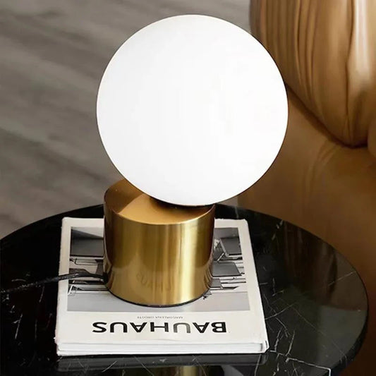 A white, spherical lamp with a soft glow sits atop a gold, cylindrical base, which rests on a book titled "BAUHAUS" on a round, dark-colored table with a marble-like pattern, with a portion of a brown leather armchair visible to the right.