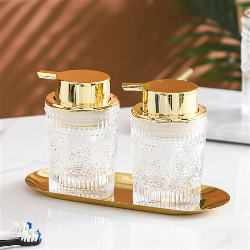 Two clear glass soap dispensers with ornate patterns and gold pumps, placed on a gold oval tray. A toothbrush and a partial view of a bathroom counter are visible.