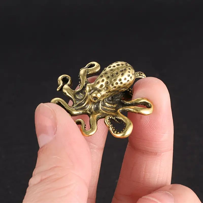 A small antique brass octopus figurine with intricate detailing, including textured tentacles and a dimpled head, held between two fingers against a dark background. The metallic finish gives it a vintage or steampunk aesthetic.