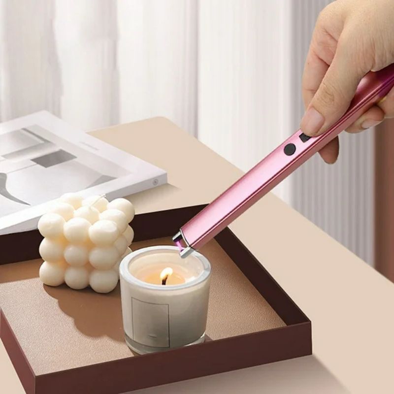 A pink electric arc lighter is being used to light a small, white candle in a glass holder. The lighter is held by a hand and positioned above the candle's wick. A white, textured decorative object is also visible in the background, along with a partially visible book or magazine. The scene is set on a brown tray.