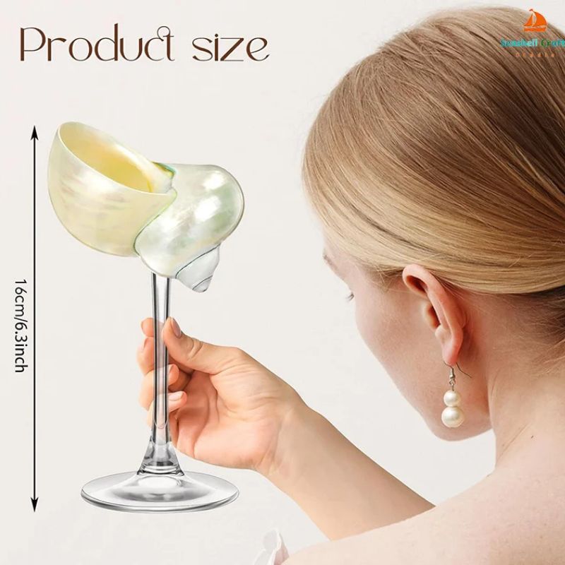 A hand holds a seashell wine glass with a clear stem and pearlescent seashell cup. The image also shows the product size of 16cm/6.3 inches in height, and a woman with light-colored hair and pearl earrings is partially visible in the background.