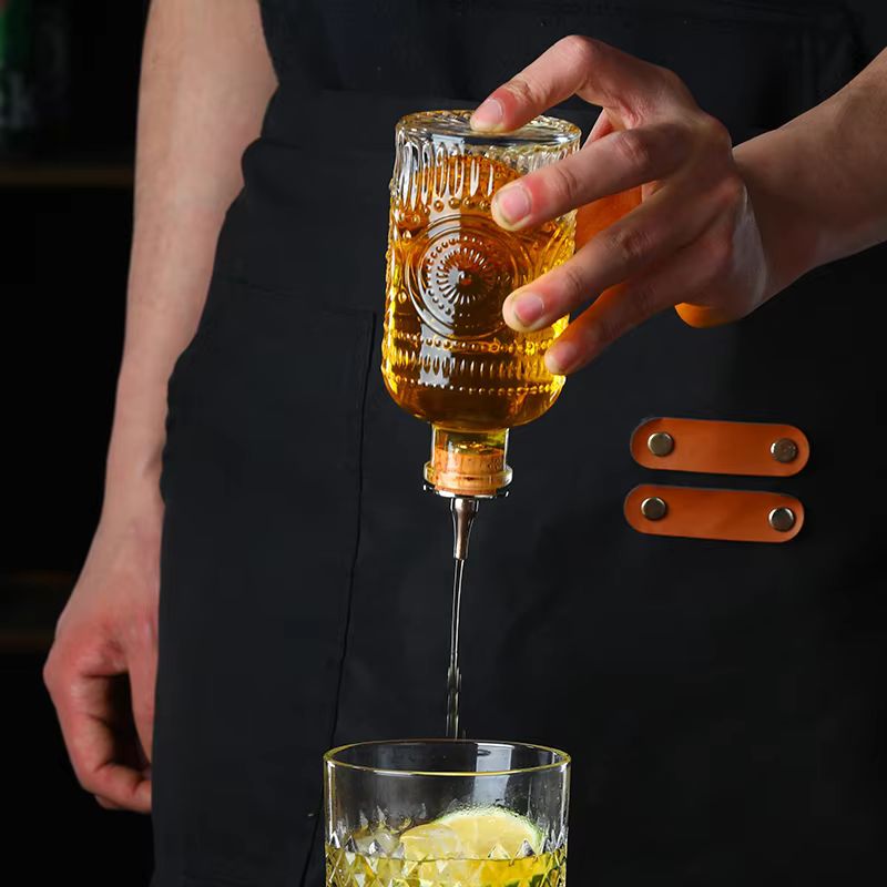 A bartender pours a golden liquid from an inverted decorative glass bottle fitted with a spout into a tumbler with ice. The bartender wears a black apron with leather strap accents.