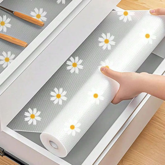 A person's hands are placing a roll of shelf liner with a white daisy pattern into a white drawer. Two wooden utensils are visible in the partially filled drawer.