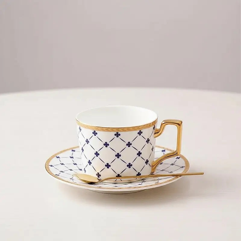 White teacup and saucer with blue lattice and clover pattern, gold trim, and a small gold spoon on a white surface.