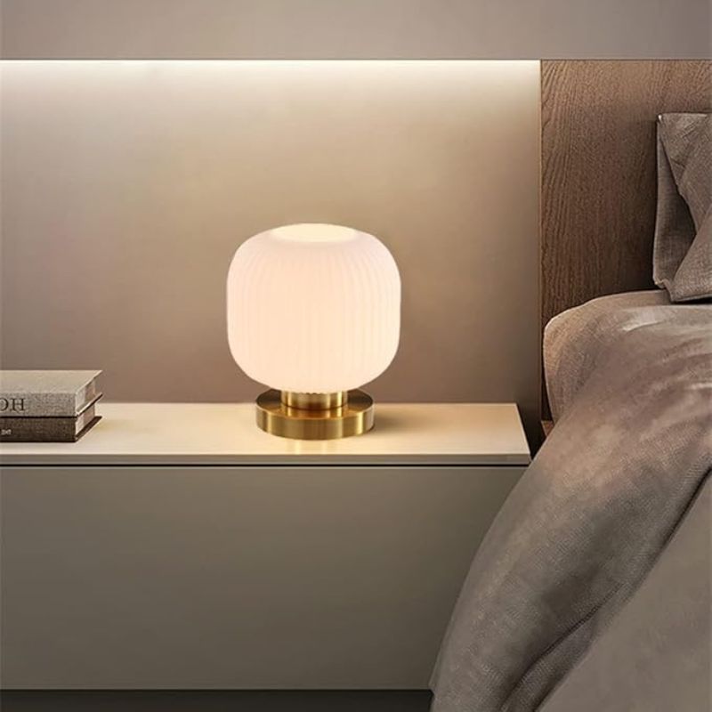A lit mid-century modern table lamp with a ribbed white glass shade and a gold base sits on a white nightstand next to a stack of books and a bed with neutral-toned bedding. A warm light strip illuminates the wall behind the nightstand.