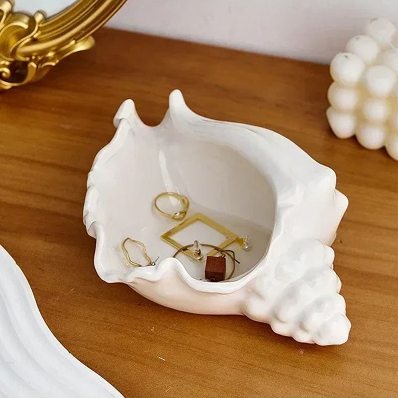 A white ceramic seashell-shaped jewelry dish placed on a wooden surface, holding gold rings and earrings, with decorative elements like a gold-framed mirror and a bubble candle in the background.
