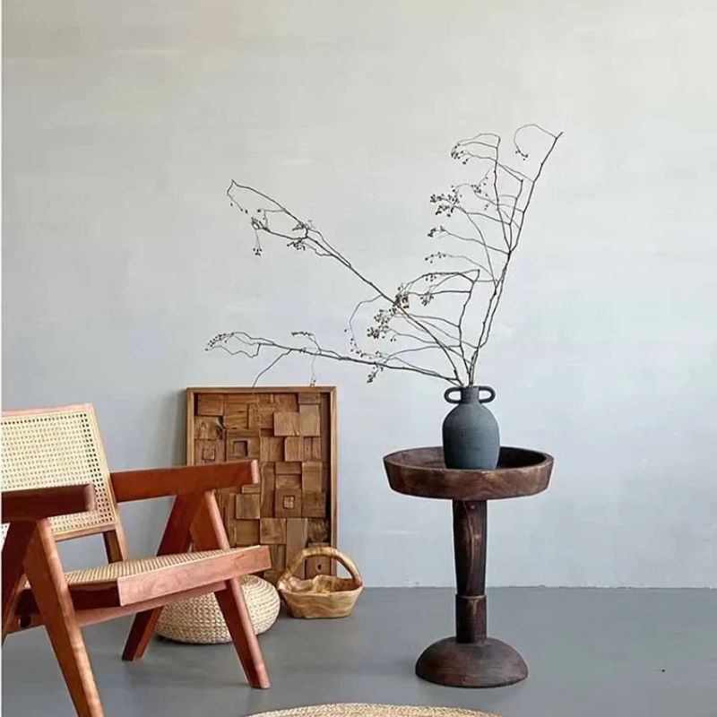 An interior scene with a dark wood, round side table holding a gray vase with bare branches, a wooden chair with a woven seat, a wooden basket, and a textured wooden wall hanging against a light gray wall.