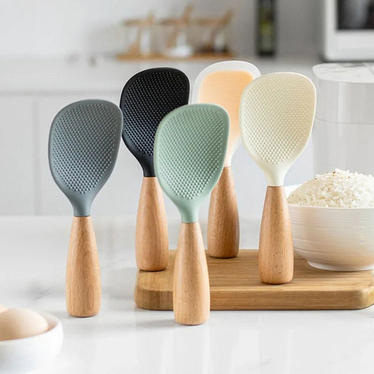 A set of five silicone and wooden rice paddles in different colors, standing upright on a wooden board, with a bowl of rice and a kitchen background.