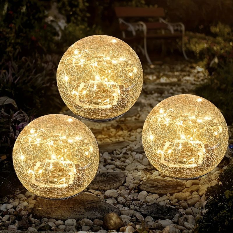 Three solar-powered crackle glass globe lights emit a warm white glow in an outdoor garden setting at night. The lights are placed on a pebble pathway, creating a cozy and decorative ambiance. A wooden bench and lush greenery are visible in the softly illuminated background.