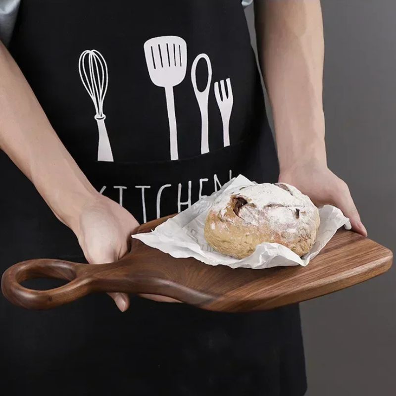 A person wearing a black apron with white kitchen utensil designs holds a small, wooden cutting board. On the board, resting on white parchment paper, is a round loaf of bread dusted with white powder. The person's hands are visible on either side of the board, and the focus is on the bread and cutting board.