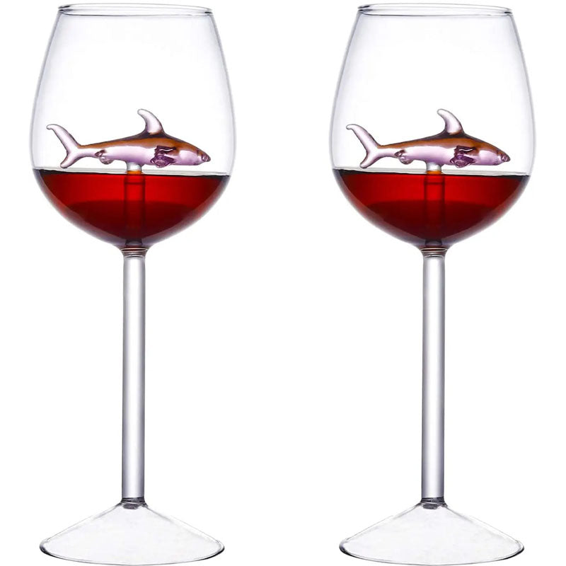 A pair of wine glasses filled with red liquid, each containing a small, pink glass shark figurine.