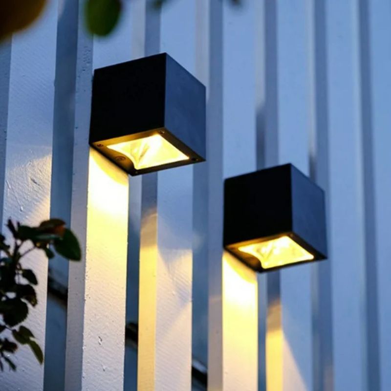 Two modern black cube-shaped outdoor wall lights mounted on a white-paneled exterior wall, emitting warm yellow light downward. The lights create an elegant and symmetrical lighting effect along the vertical panels. A few green leaves are visible in the foreground, adding a natural element to the composition.