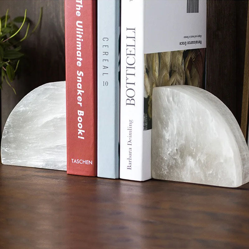 A pair of white semi-circular crystal bookends holding upright books on a wooden surface, with a small green plant in the background.