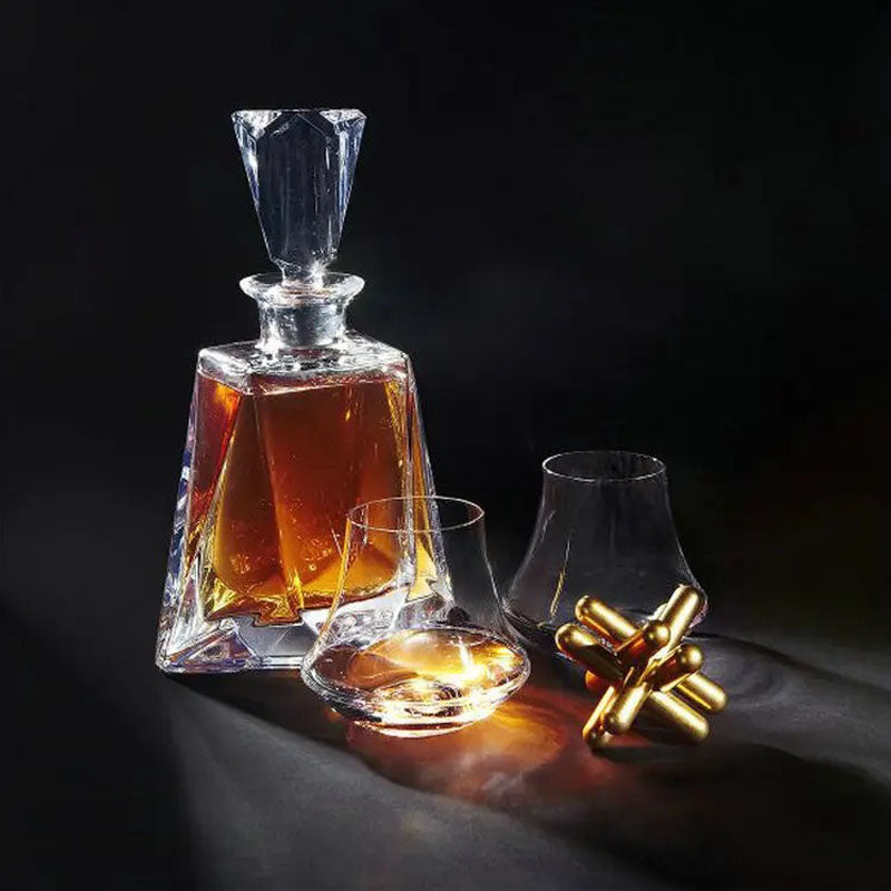A geometric whiskey decanter filled with amber liquid, accompanied by two uniquely shaped whiskey glasses and a metallic gold decorative piece, all set against a dramatic dark background.