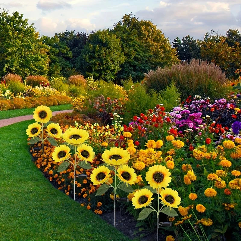 A vibrant garden features a curved row of artificial sunflowers with dark centers and bright yellow petals lining a green lawn. Beyond the sunflowers, a colorful array of real flowers in shades of yellow, orange, red, pink, and purple blooms in a lush flowerbed. A winding dirt path cuts through the garden, leading towards mature trees and varied greenery under a partly cloudy sky.