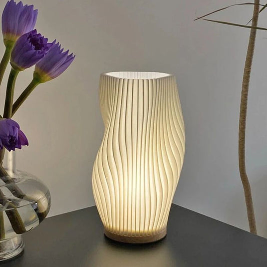 Modern table lamp with a sculptural, wavy design, emitting a warm glow. Placed on a dark surface beside a glass vase with purple flowers.