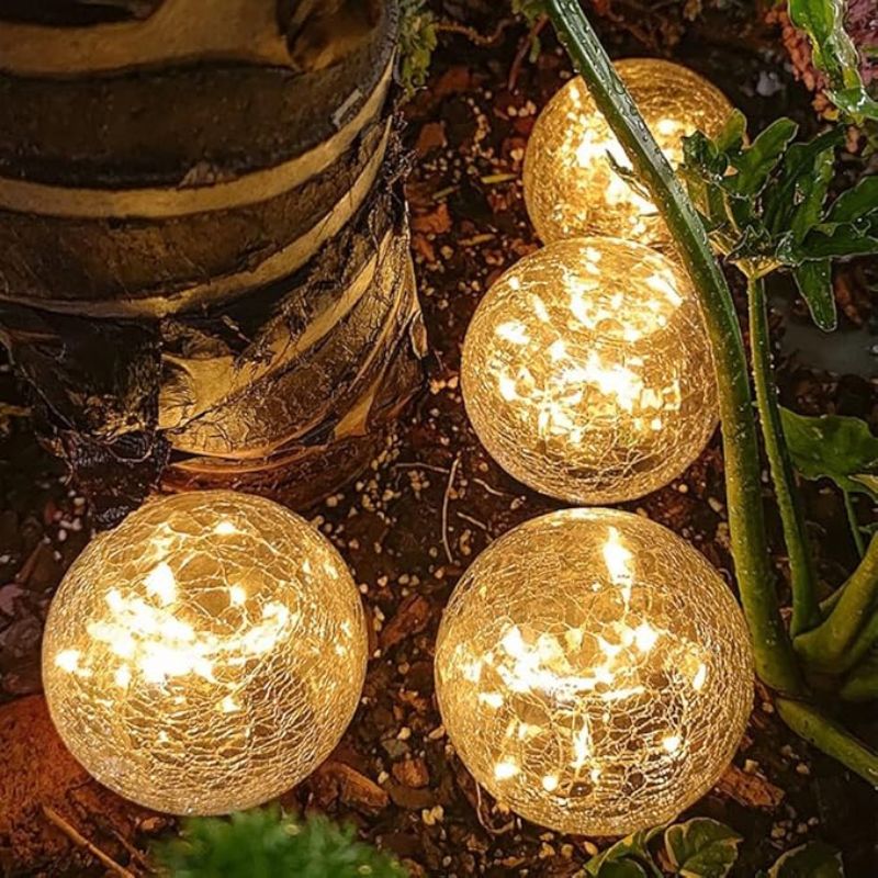 Four crackle glass solar garden lights emitting a warm golden glow, placed on the ground near a tree and surrounded by green plants. The lights create a cozy and decorative ambiance in the outdoor setting.