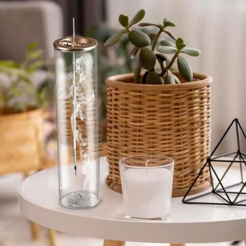 This image features a cozy indoor setting with a white round table. On the table, there's a tall cylindrical glass incense holder with a gold-colored lid, where an incense stick is burning, releasing delicate swirls of smoke. Next to it, there's a small glass-encased white candle. In the background, a woven rattan planter holds a green succulent plant, and a black geometric metal ornament rests beside it. The setting has a warm, minimalist, and natural aesthetic.