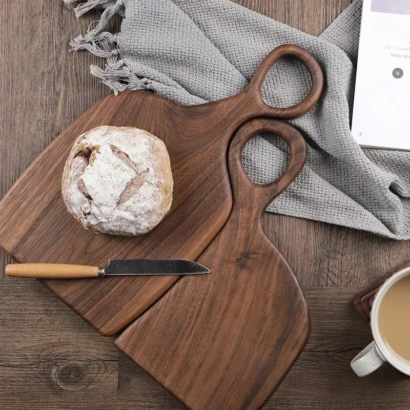 Two wooden cutting boards, one with a round loaf of bread and a knife on it, are arranged on a dark wooden surface with a gray cloth, a book, and a mug of coffee.