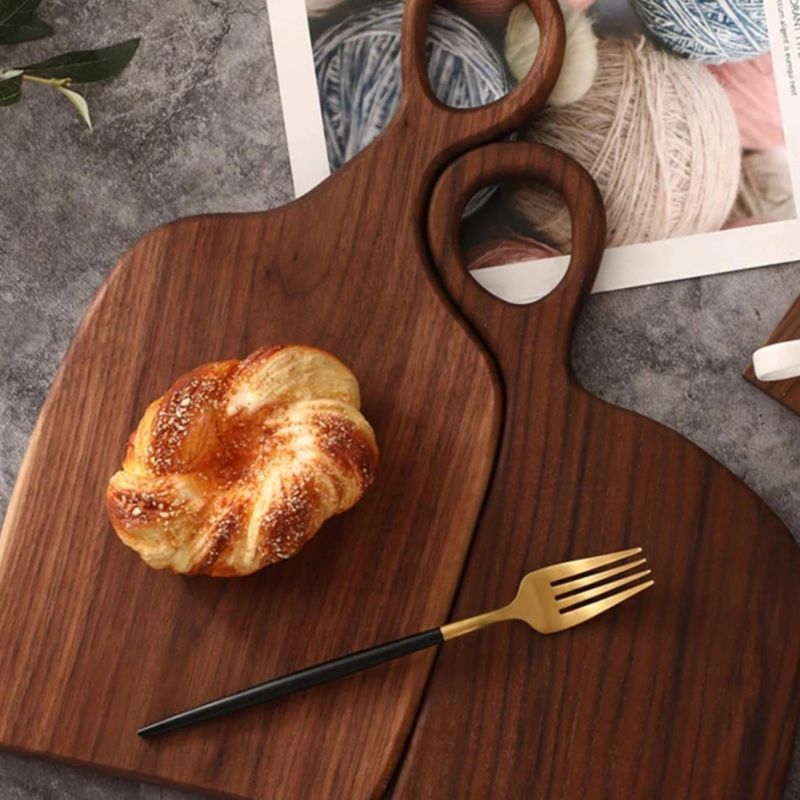 Two wooden cutting boards with circular handles are arranged on a textured gray surface. A golden-brown pastry sits on the smaller board, while a gold and black fork rests on the larger one. A magazine or book showing yarn is partially visible in the upper right.