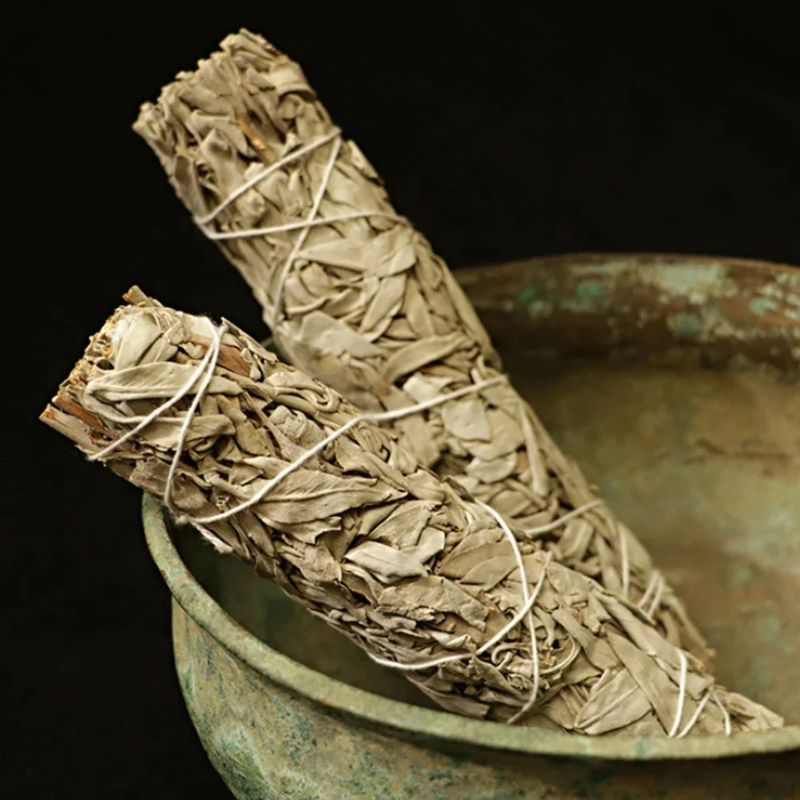 A close-up shot of two bundles of dried white sage, tied with white string, resting inside a rustic, weathered bronze bowl. The sage bundles are the primary focus, showing their characteristic pale gray-green leaves and rough texture. The bowl appears aged with a greenish patina and a slightly uneven rim, suggesting a vintage or handcrafted piece. The background is a dark, solid color, providing a stark contrast that highlights the sage and the bowl.