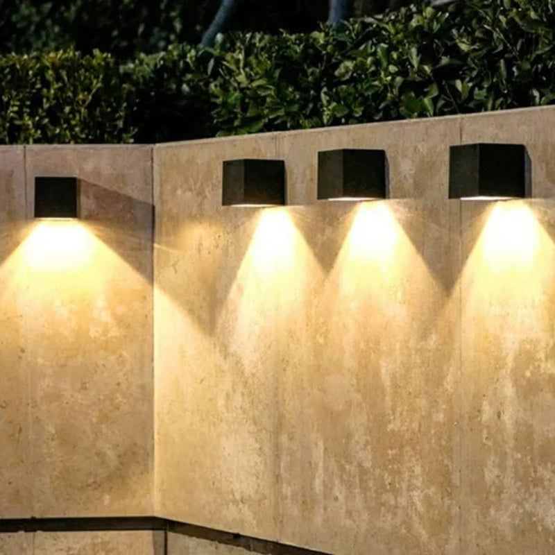 Four modern black cube-shaped outdoor wall lights mounted on a beige stone wall, casting a warm downward glow. The lights are evenly spaced, creating a visually appealing and symmetrical illumination effect. Green foliage is visible at the top of the image, adding a natural contrast to the structured lighting.