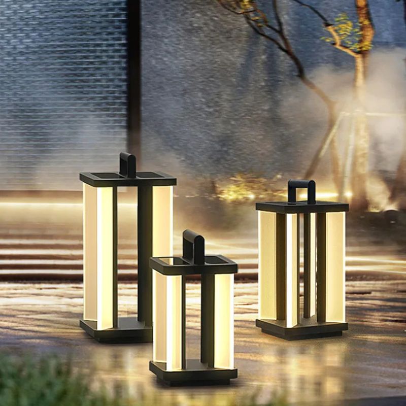 Three illuminated "Villa Solar Lamps" of varying sizes stand on a textured outdoor surface near a building with reflective glass and a bare tree.