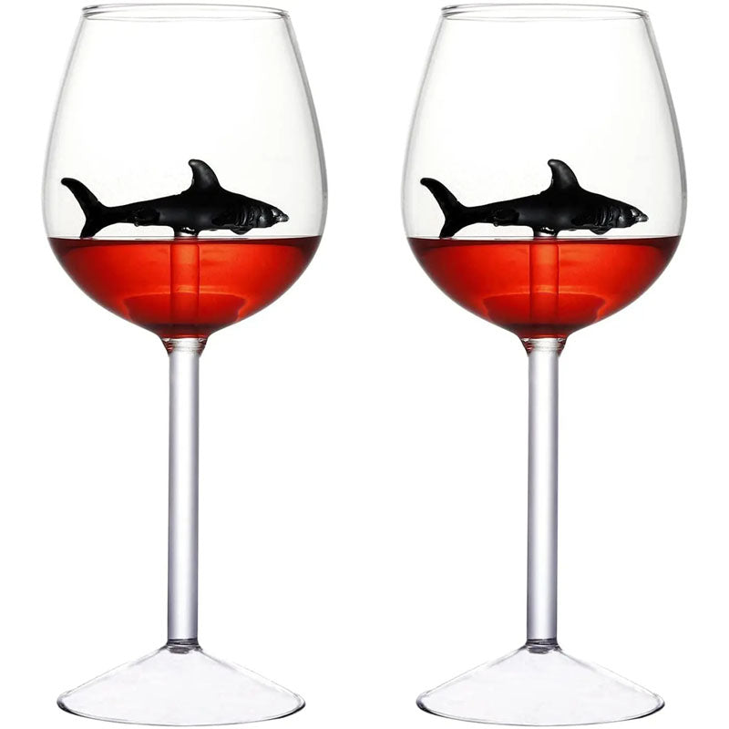 A pair of wine glasses filled with red liquid, each containing a small, black glass shark figurine.