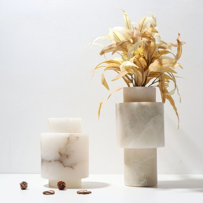 Two Scandinavian White Onyx vases displayed on a white surface against a light background. The taller vase elegantly holds dried golden foliage, while the shorter vase showcases natural marble veining. Small pinecones and dried leaves are scattered around, adding a warm, organic touch to the modern aesthetic.