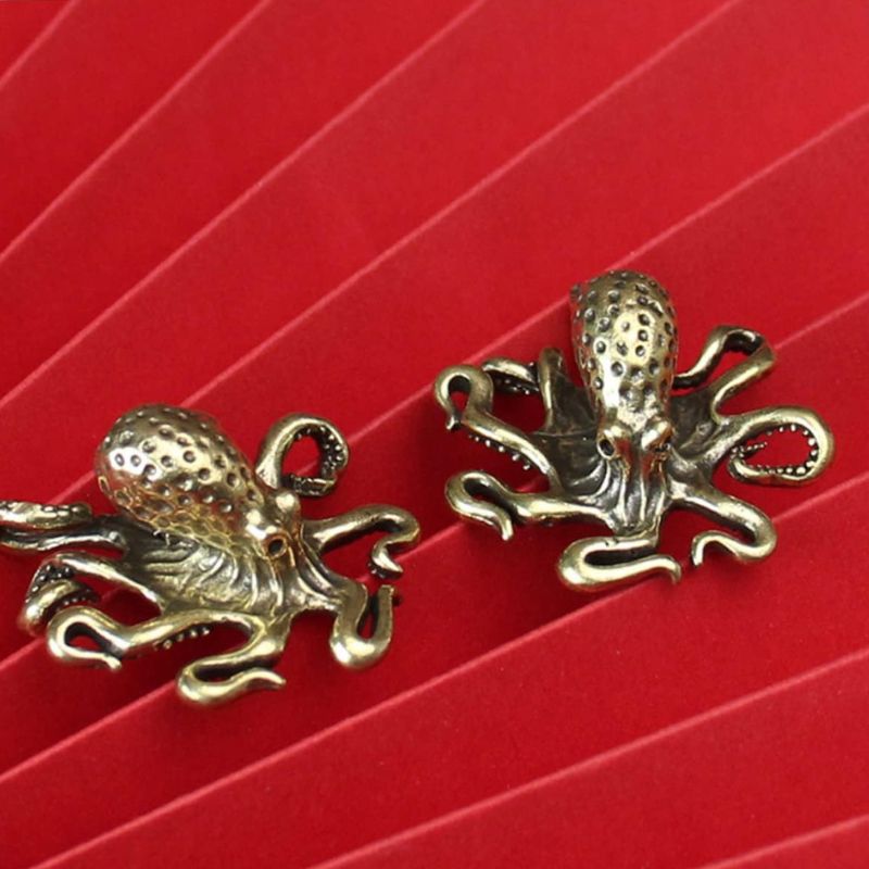 A pair of antique brass octopus figurines with intricate detailing, including curled tentacles and a textured head, placed on a red folded paper background. The metallic finish gives them a vintage or steampunk aesthetic.