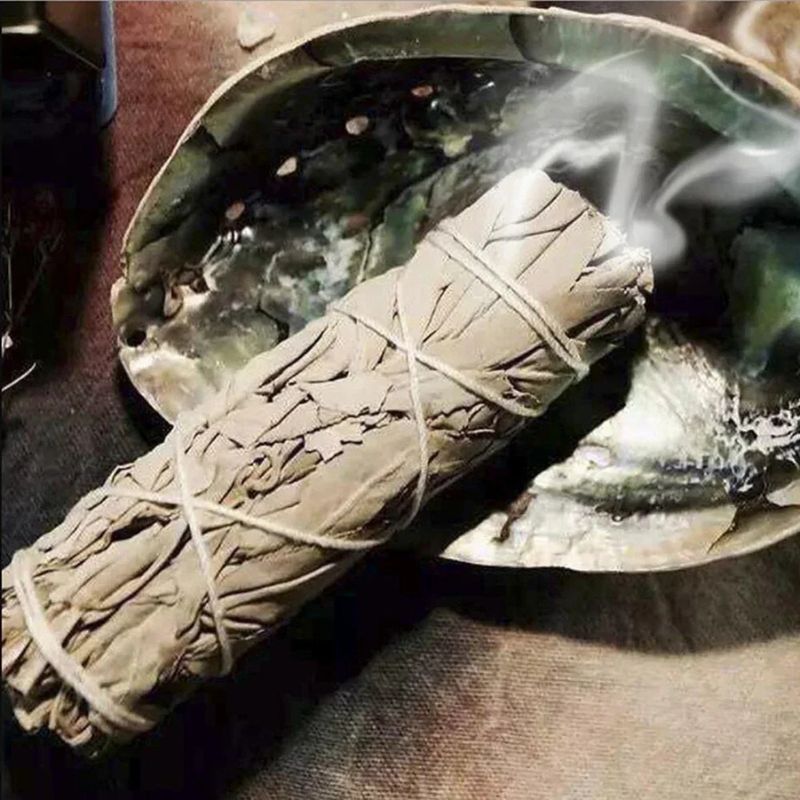 A close-up shot of a thick bundle of dried white sage, tied with white string, resting in a large, iridescent abalone shell. The sage bundle is the primary focus, showing its pale gray-green leaves and rough texture. Wisps of white smoke rise from the sage, indicating it is being burned. The abalone shell has a pearlescent sheen with shades of green, blue, and silver, and its interior is visible, adding to the visual interest. The background is a textured, dark surface, providing contrast and highlighting t