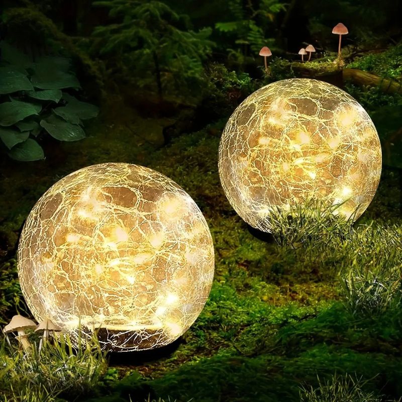 Two solar-powered crackle glass globe lights emit a warm white glow in a lush, moss-covered garden at night. The lights are nestled among greenery, with small mushrooms growing nearby, creating an enchanting and magical outdoor ambiance.