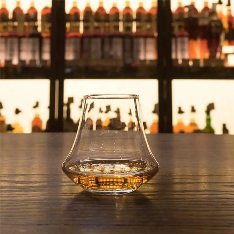 A modern whiskey glass with a sleek, angular design, partially filled with golden liquor, set against a warmly lit bar backdrop.