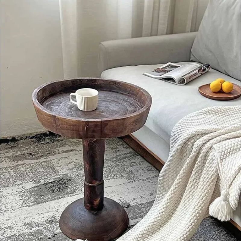 A rustic, dark wood, round side table (Momo Wabi-sabi Log Coffee Table) with a shallow tray top, holding a white cup, positioned next to a light-colored sofa with a magazine, lemons, and a white blanket.