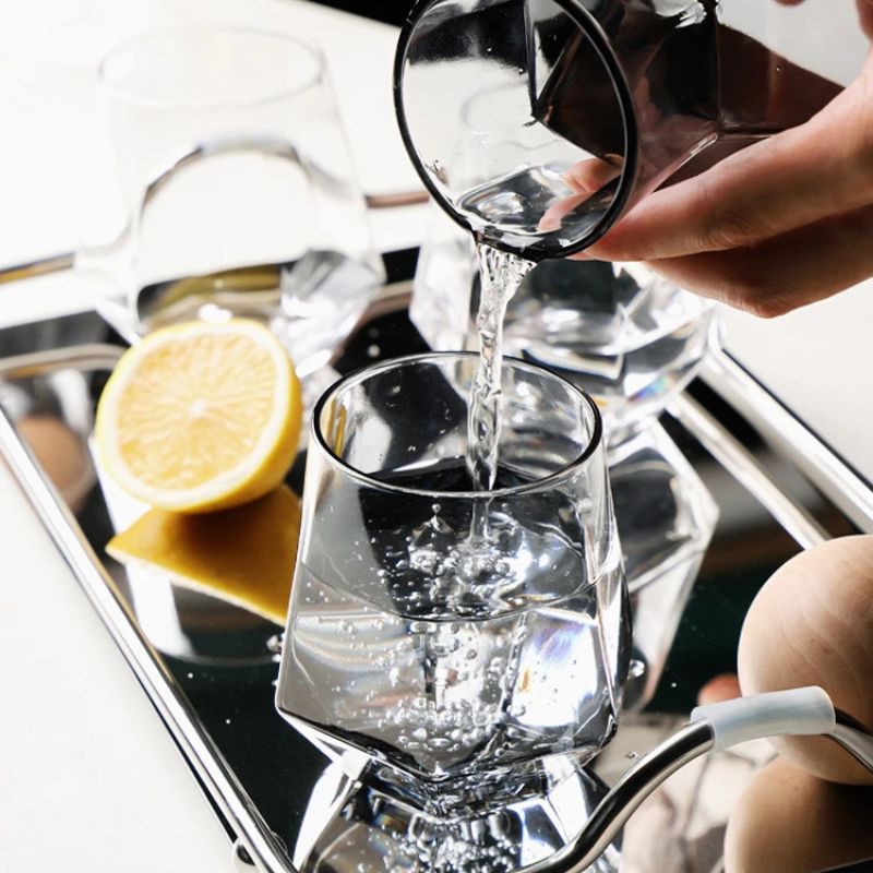 Close-up of a hand pouring water from a dark gray faceted glass into a clear faceted glass on a silver tray, with a lemon slice and a wooden object visible.