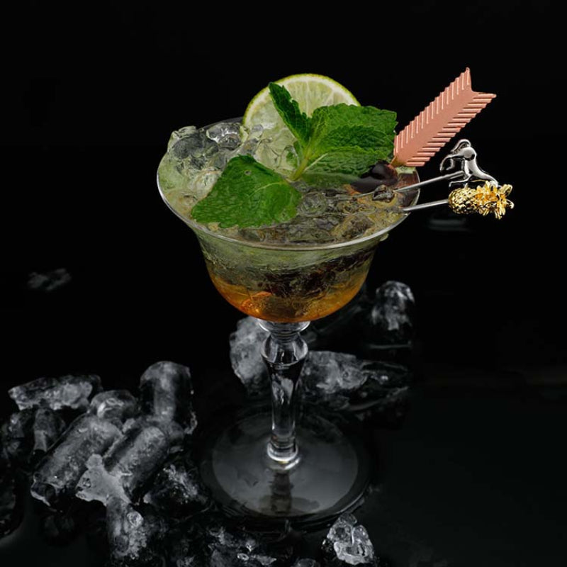 A cocktail in a stemmed glass with mint leaves, lime, and decorative cocktail picks, surrounded by ice on a black background.