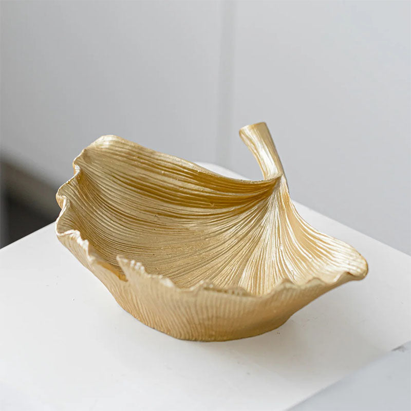 Gold-colored decorative bowl with a sculpted, textured design resembling a ginkgo leaf, placed on a white surface.