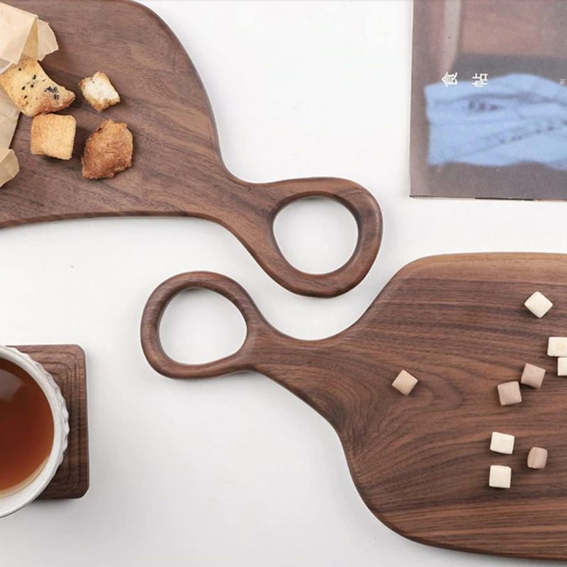 Two wooden cutting boards with circular handles are arranged on a dark surface. The top board holds several small, irregularly shaped crackers in a partially open brown paper bag. The bottom board is scattered with small, white square candies. A white mug with a light-colored beverage sits on a matching wooden coaster in the lower left corner. A book or magazine with text in a non-English script is partially visible in the upper right.