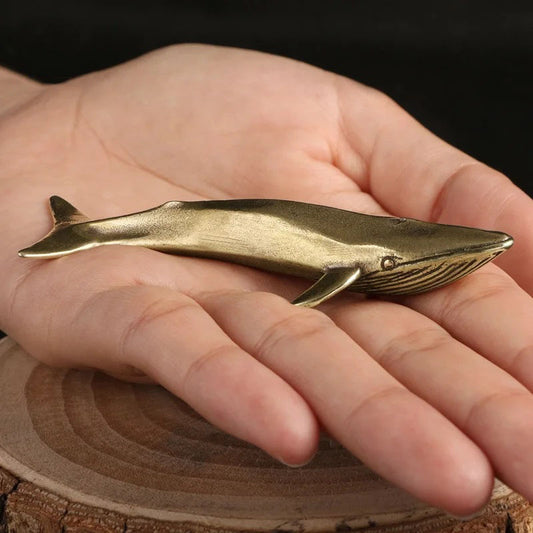 A small, brass-colored whale figurine is held in a person's open palm. The figurine is positioned horizontally across the palm, and the palm is cupped slightly. The background is a dark surface.