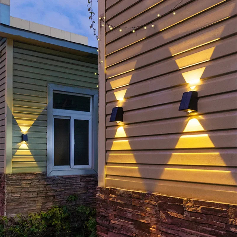 Three small, dark, rectangular lights are mounted on the exterior walls of two adjacent buildings with horizontal siding and brick bases. Each light casts a warm white triangular beam both upwards and downwards at dusk or night. String lights are visible above.