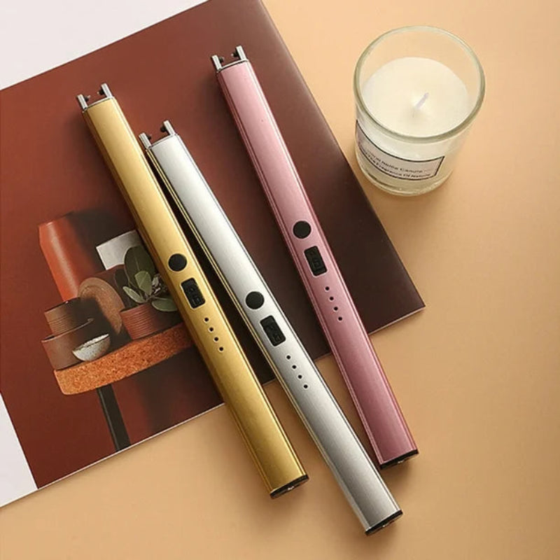 Three slim, cylindrical electric arc lighters in gold, silver, and pink, arranged diagonally on a light brown surface with a magazine or booklet and a small glass candle holder in the background.