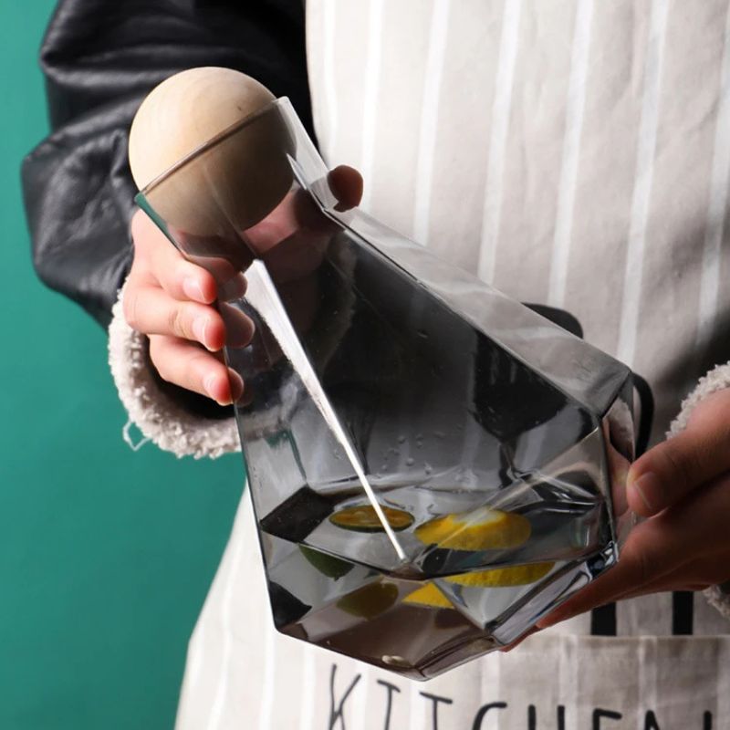Hands holding a dark gray faceted glass pitcher with a wooden ball lid, containing water and lemon slices. A striped apron and part of a dark jacket are visible in the background.