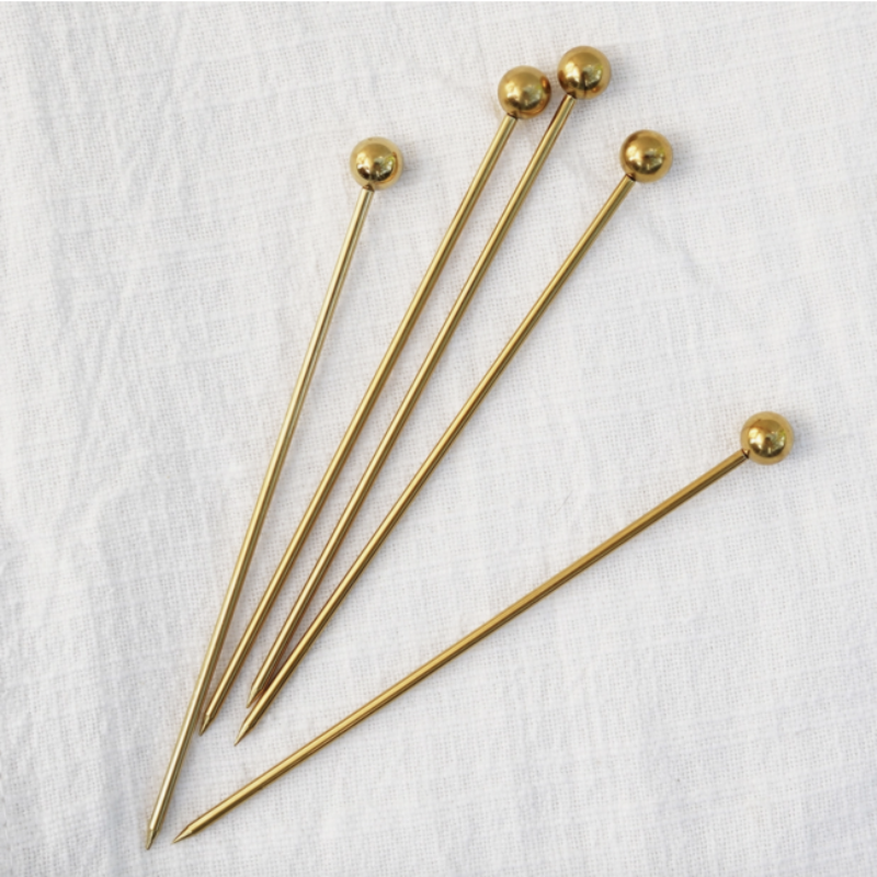 Five long, thin gold cocktail picks with small, smooth spherical ends, fanned out on a white fabric surface with a subtle woven texture.